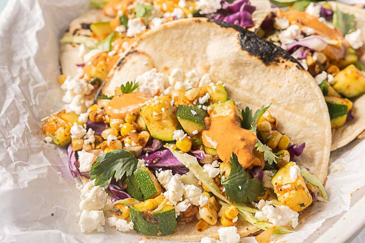 Three corn tortillas filled with grilled zucchini, corn, shredded purple cabbage, cilantro, crumbled cheese, and an orange sauce are arranged on parchment paper—a perfect pick for summer recipes or creative zucchini recipes.