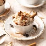 A white ramekin filled with almond milk rice pudding, garnished with raisins, almond slivers, and a cinnamon stick, placed on a white plate. Other ramekins are visible in the background.