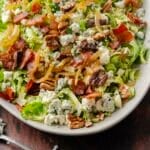 A bacon brussels sprout salad with mixed greens, blue cheese crumbles, candied nuts, caramelized onions, and crispy bacon pieces is beautifully presented on a white plate, with a fork on the side.
