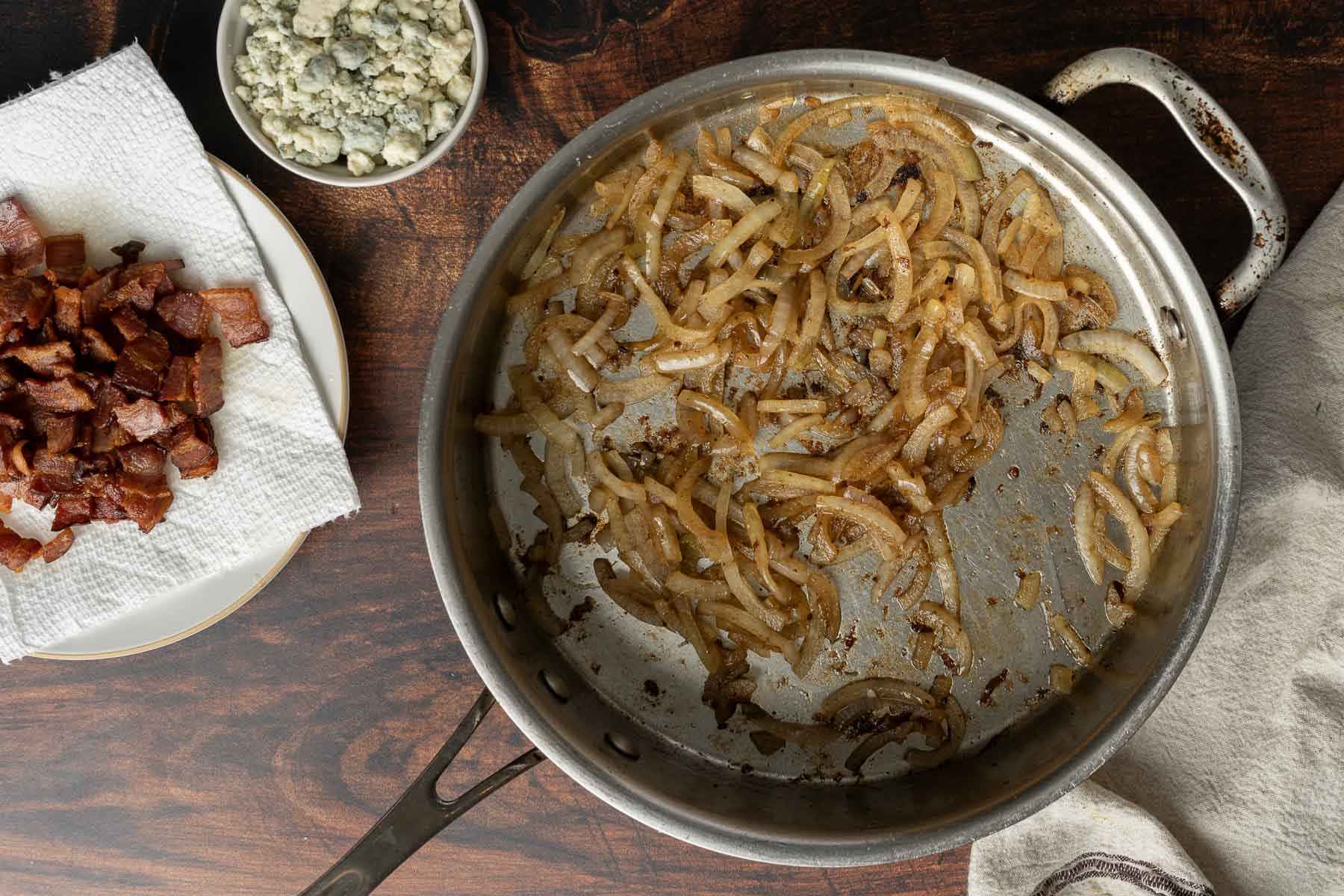 A skillet with cooked onions sits on a wooden surface. Nearby, a plate with crispy bacon on a paper towel and a bowl of crumbled blue cheese are visible, setting the stage for a delicious bacon Brussels sprout salad.