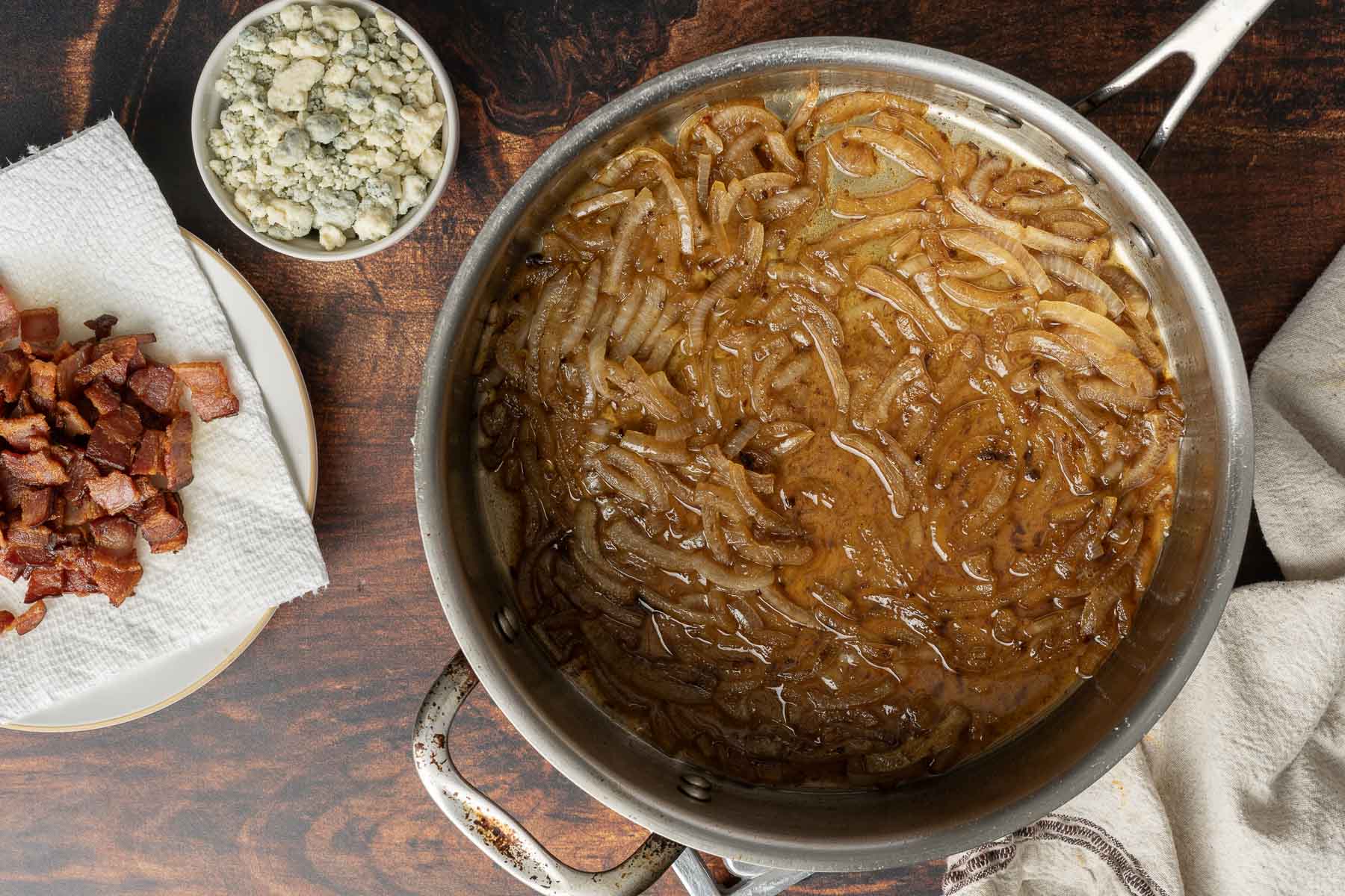 A skillet filled with caramelized onions beside a plate with cooked bacon pieces on a paper towel and a bowl of crumbled blue cheese, all ready to be mixed into a mouthwatering bacon Brussels sprout salad.