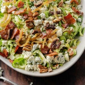 A bacon brussels sprout salad with mixed greens, blue cheese crumbles, candied nuts, caramelized onions, and crispy bacon pieces is beautifully presented on a white plate, with a fork on the side.