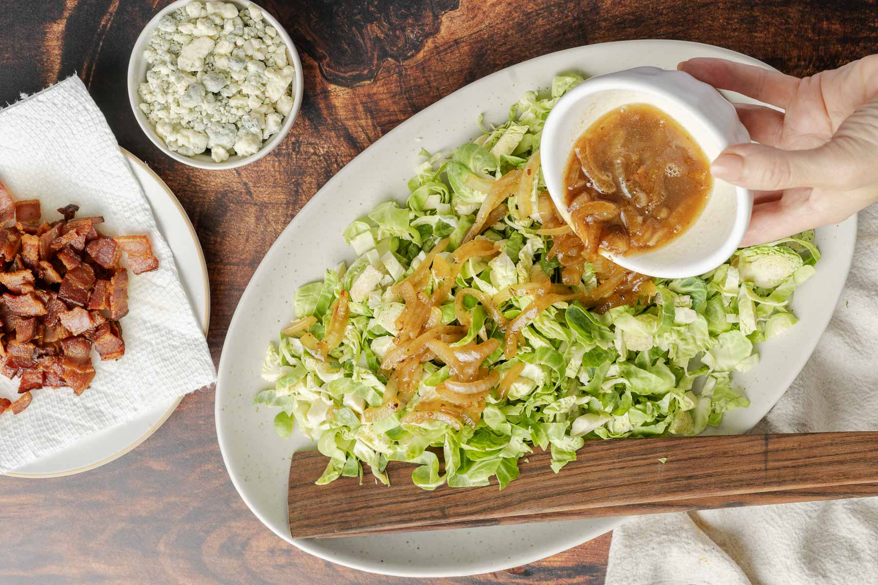 A hand pours dressing onto a chopped bacon Brussels sprout salad on an oval plate. Next to the plate are a small bowl of blue cheese crumbles and a dish with chopped cooked bacon.