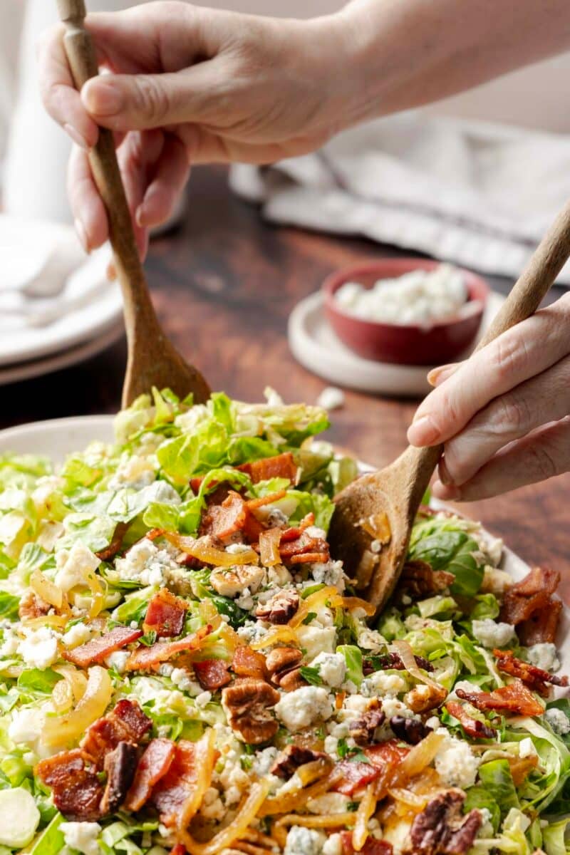Close-up of hands using wooden utensils to toss a bacon brussels sprout salad with mixed greens, blue cheese, nuts, and caramelized onions.