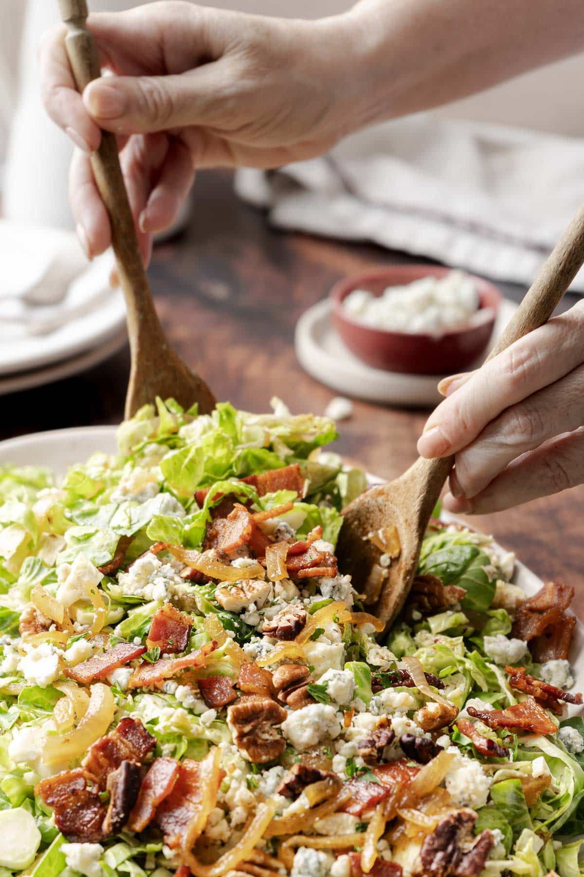 Close-up of hands using wooden utensils to toss a bacon brussels sprout salad with mixed greens, blue cheese, nuts, and caramelized onions.