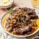 A plate of oven-baked lamb shoulder chops garnished with caramelized onions and fresh thyme. A bowl of mashed potatoes and a glass of yellow liquid are in the background.