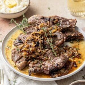 A plate of oven-baked lamb shoulder chops garnished with caramelized onions and fresh thyme. A bowl of mashed potatoes and a glass of yellow liquid are in the background.