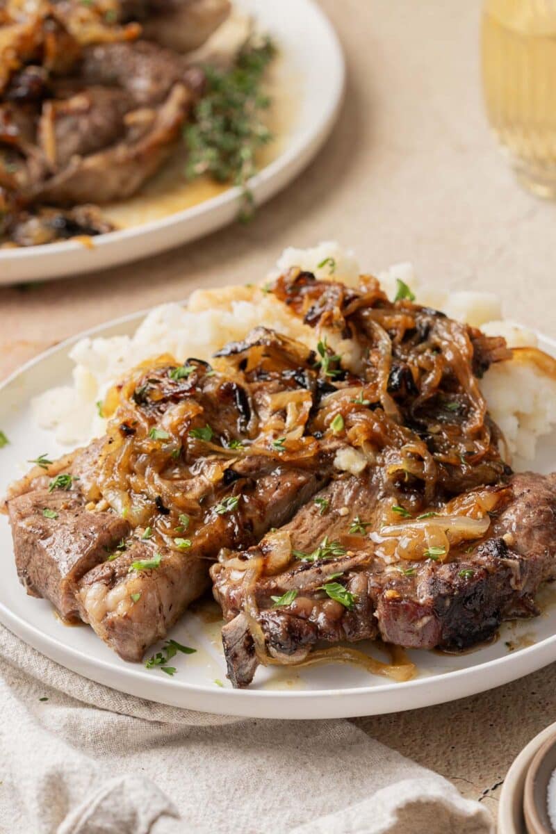 A plate of oven-baked lamb shoulder chops topped with caramelized onions, garnished with herbs, and served with mashed potatoes, set on a beige tablecloth.