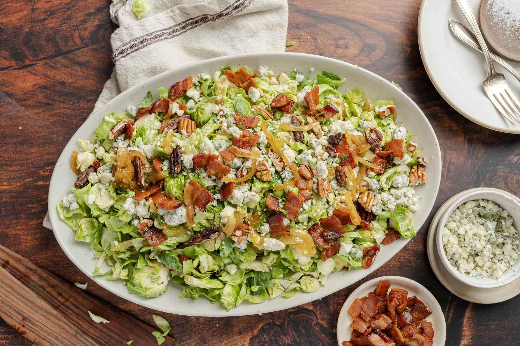A platter of bacon brussels sprout salad featuring crumbled blue cheese, pecans, and caramelized onions sits on a wooden table. A side of extra blue cheese and bacon accompanies the dish, with plates and a napkin nearby.