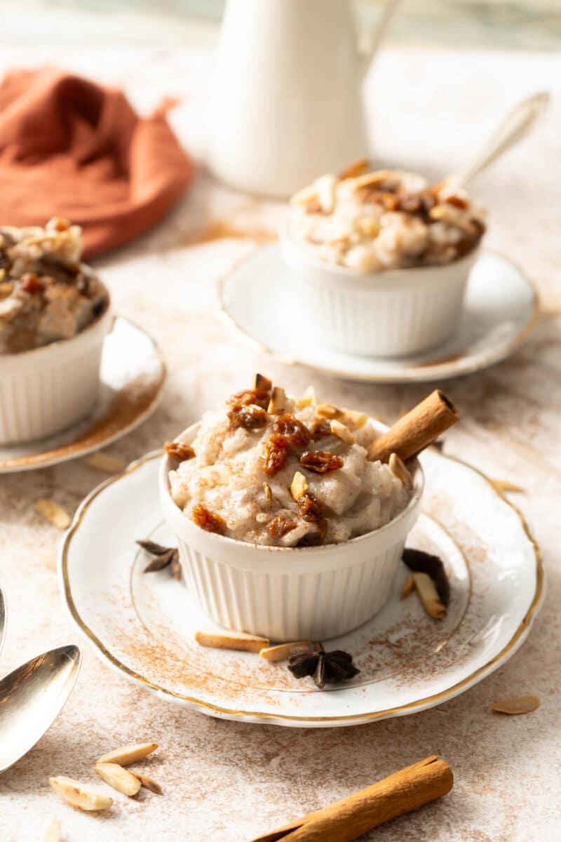 Three small dishes of almond milk rice pudding topped with nuts and a cinnamon stick, served on white plates. A jug, scattered nuts, and a spoon are visible in the background.