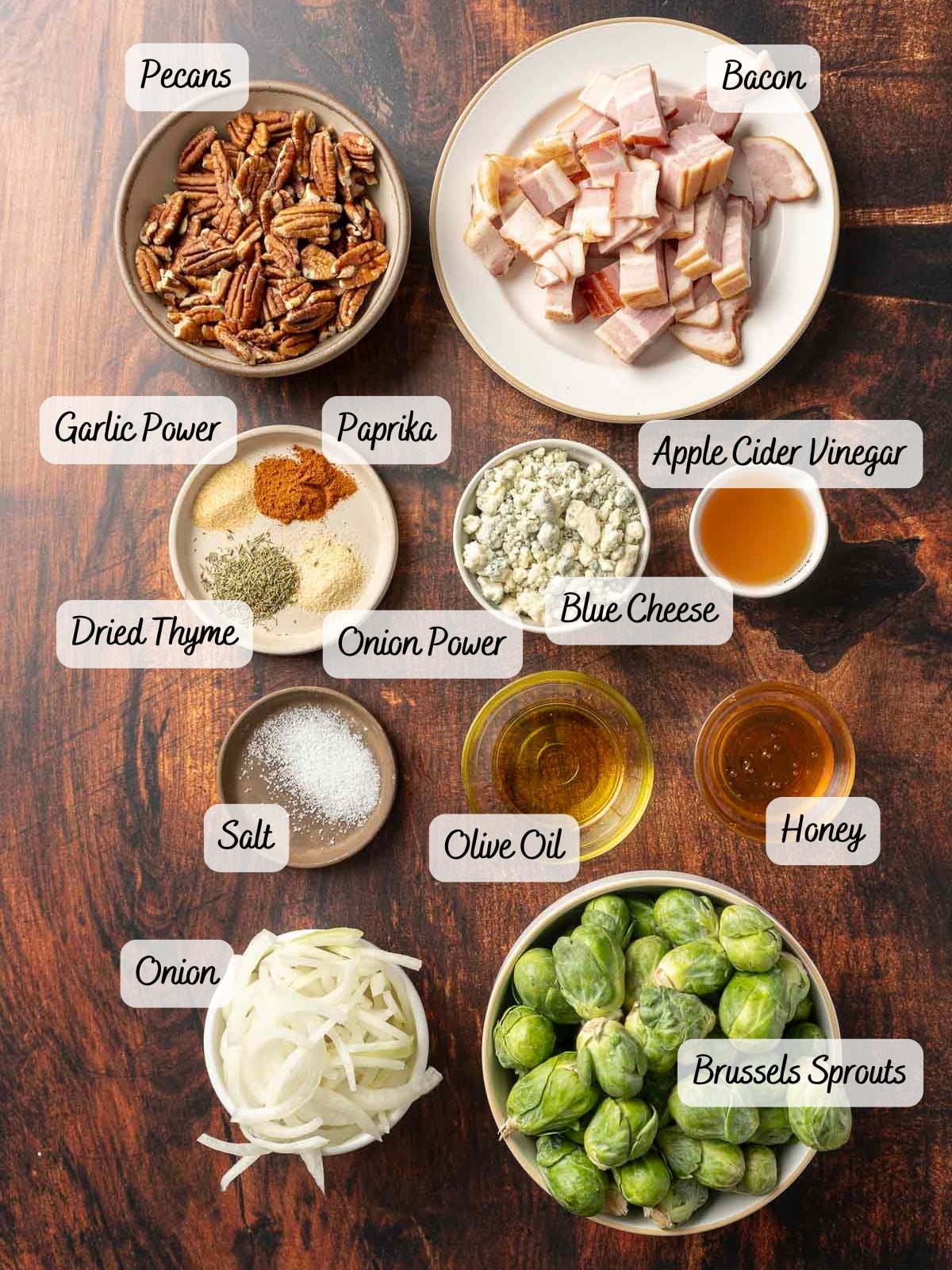 Ingredients on a table include a bowl of pecans, garlic powder, paprika, apple cider vinegar, dried thyme, blue cheese, onion powder, honey, olive oil, salt, sliced onion and Brussels sprouts for a delicious bacon Brussels sprout salad.