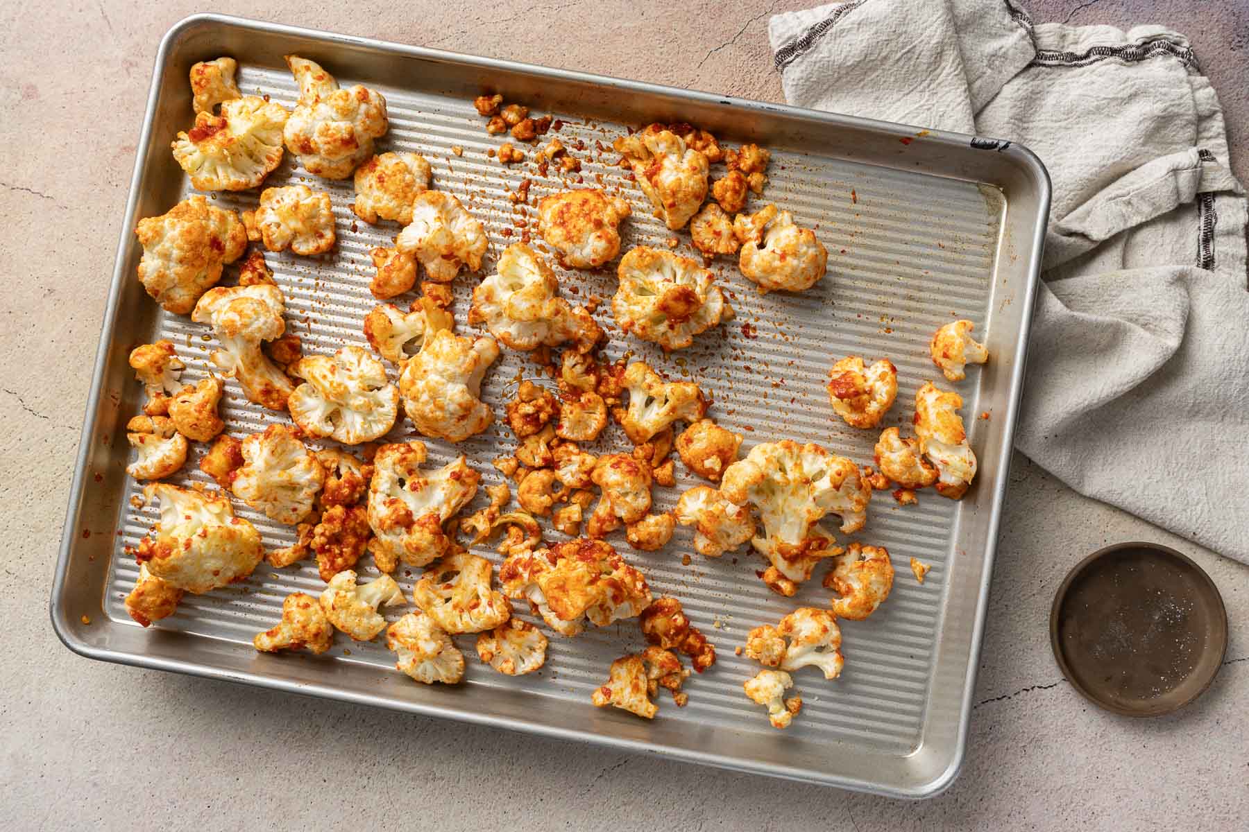 A baking sheet with harissa cauliflower florets, seasoned and slightly browned. An off-white cloth napkin and an empty dish are placed beside the tray.