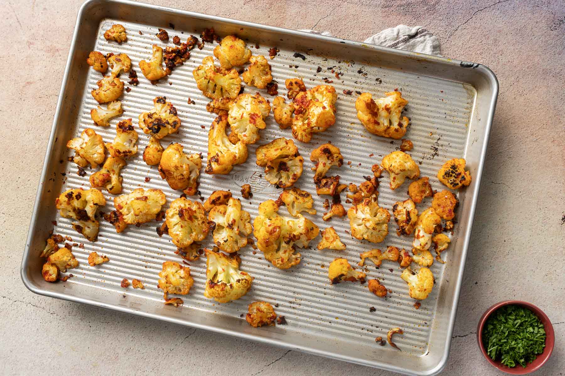 A baking sheet with harissa cauliflower florets, evenly spread and lightly browned. A small bowl of chopped herbs sits beside the sheet.