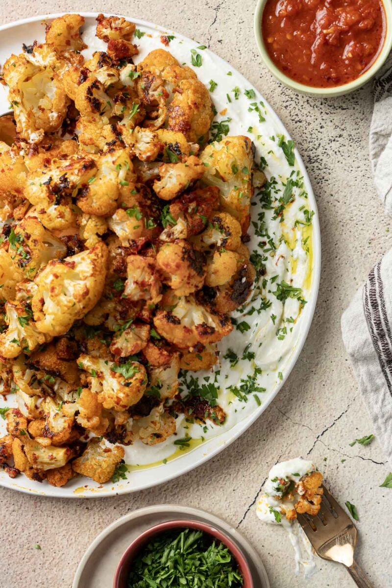 A plate of harissa cauliflower garnished with fresh herbs, resting on a sauce, with small bowls of dipping sauce and chopped herbs on the side. A fork with a small piece of cauliflower is nearby.