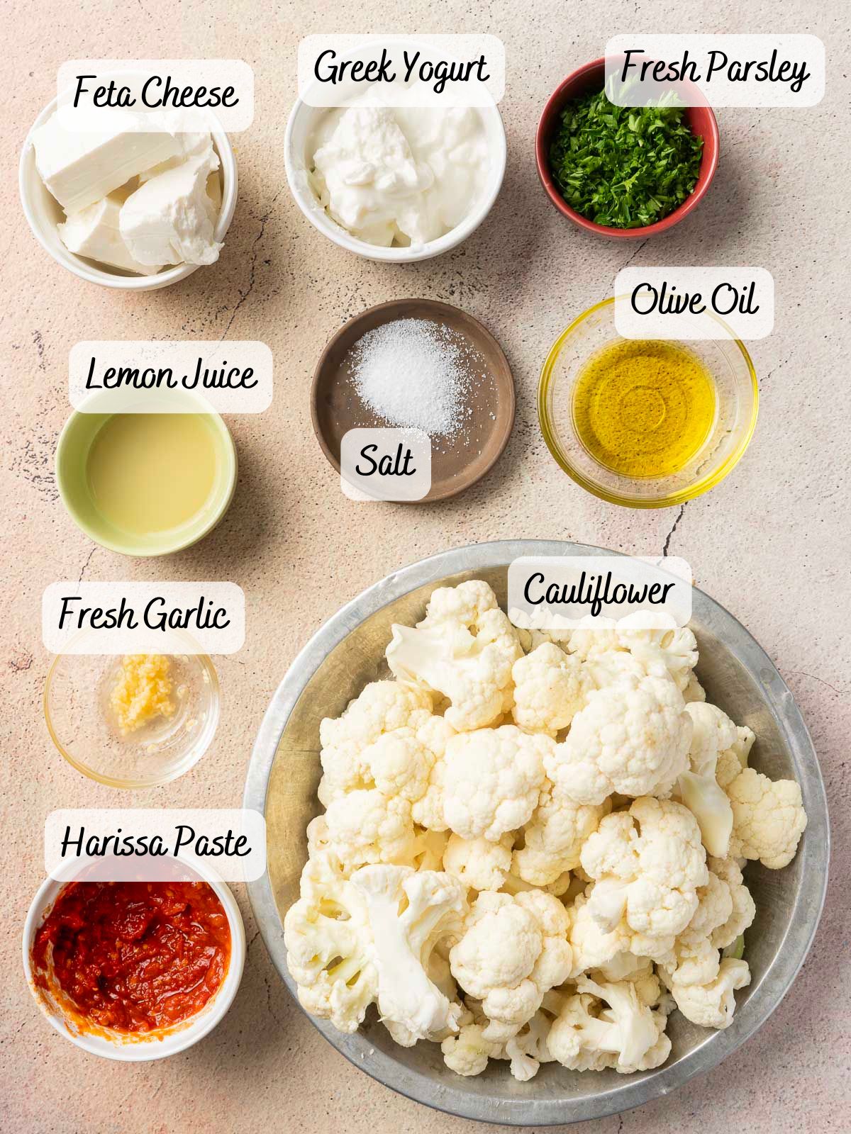 Top view of ingredients arranged in bowls and identified with labels: Greek yogurt, feta cheese, fresh parsley, lemon juice, salt, olive oil, fresh garlic, harissa cauliflower florets.