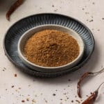 A bowl of ground harissa seasoning is placed inside a larger dark blue plate, with dried chilies and scattered spice fragments visible in the background.