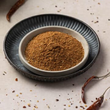 A bowl of ground harissa seasoning is placed inside a larger dark blue plate, with dried chilies and scattered spice fragments visible in the background.