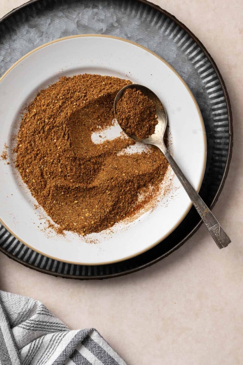 A white plate with a heaping spoon of brown ground spices, likely a harissa seasoning blend, is placed on a textured black plate. A striped cloth is partially visible at the bottom left corner.