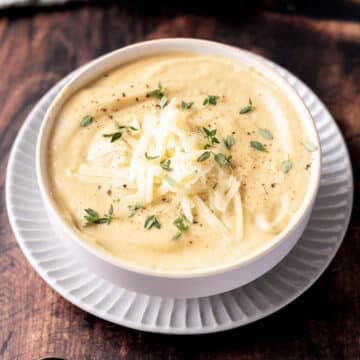 A bowl of creamy rutabaga soup garnished with shredded cheese, fresh herbs, and black pepper, served on a patterned white plate on a wooden table.