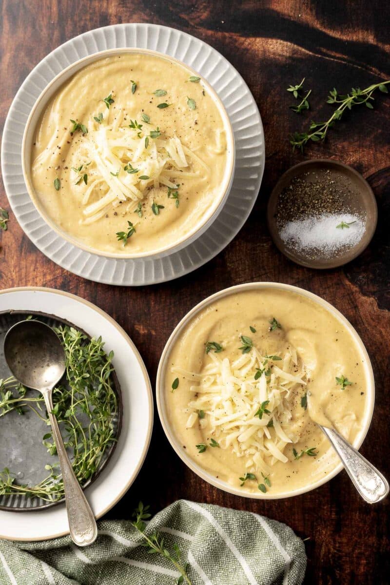 Two bowls of creamy rutabaga soup garnished with shredded cheese and herbs, accompanied by spoons, a dish with a salt and pepper mix, fresh herbs, and a green cloth napkin on a wooden table.