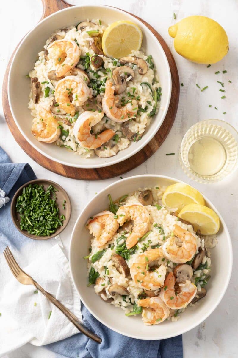 Two bowls of shrimp risotto garnished with lemon slices and chives, accompanied by a small bowl of chives, a glass of white wine, and a blue napkin with a fork on a white surface.