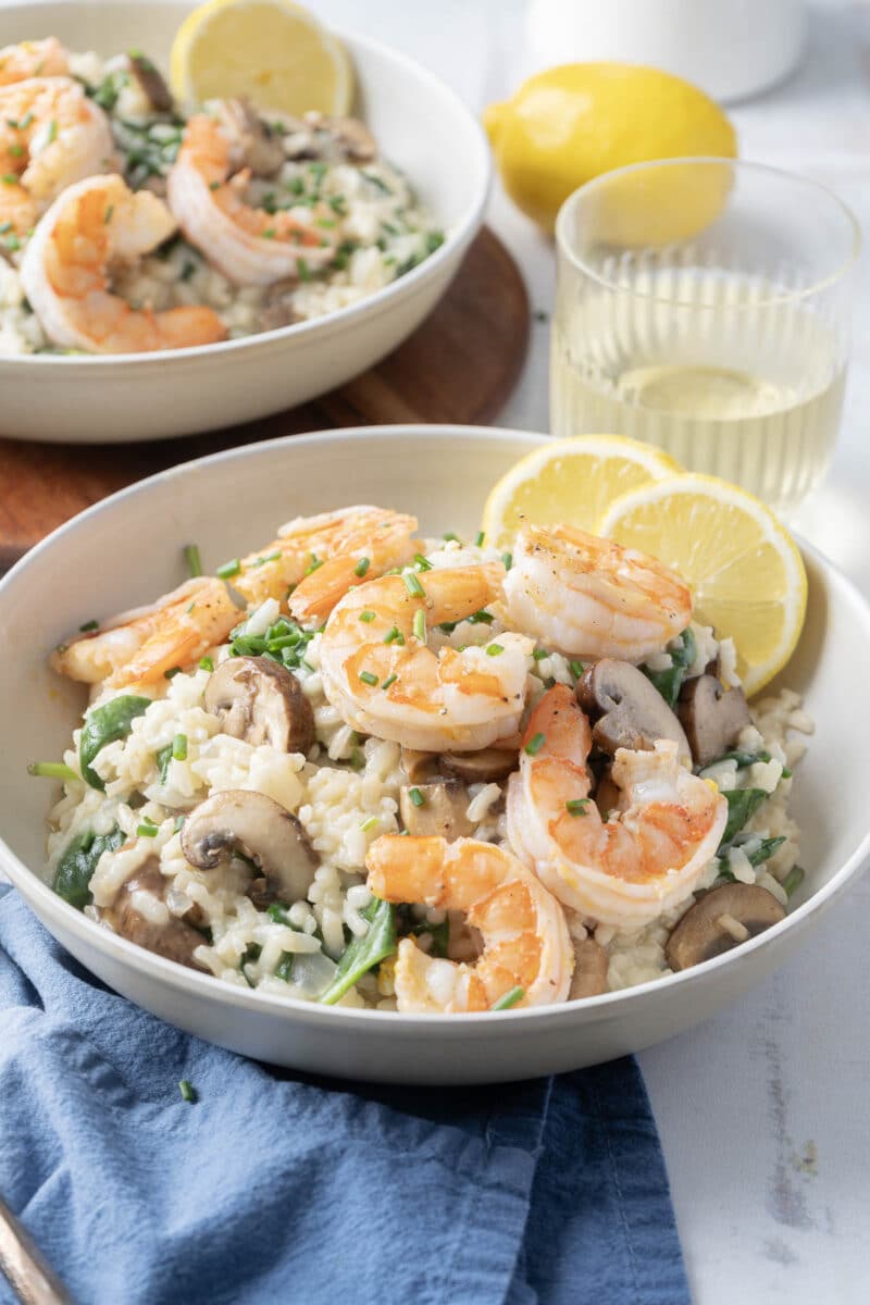 A bowl of shrimp risotto garnished with lemon slices and chives, alongside spinach. A glass of white wine and another bowl of risotto are in the background, with a blue napkin nearby.