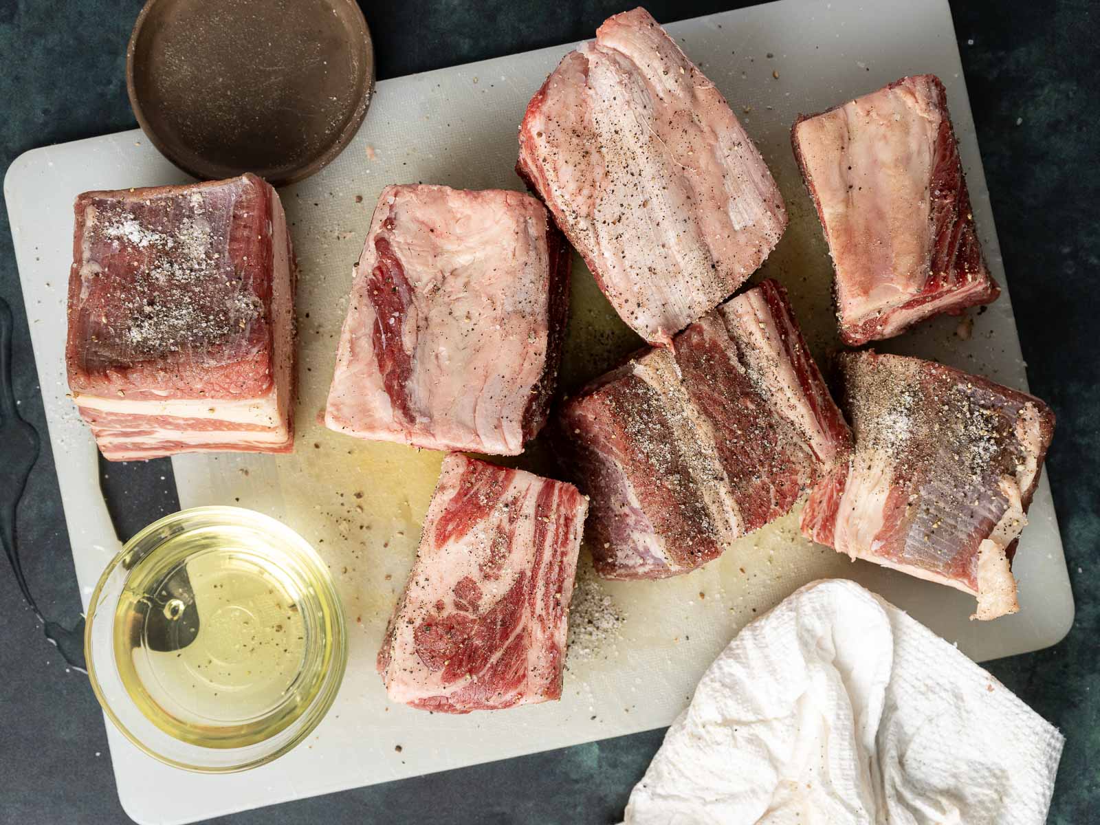 Raw beef short ribs with seasoning rest on a cutting board, accompanied by a bowl of oil and a white cloth, ready to transform into a rich braised beef ragu.