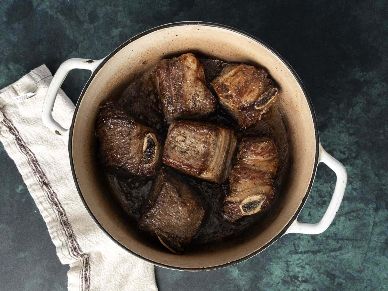 A pot of tender braised beef short ribs rests on a dark surface, with a white cloth elegantly placed beside it.