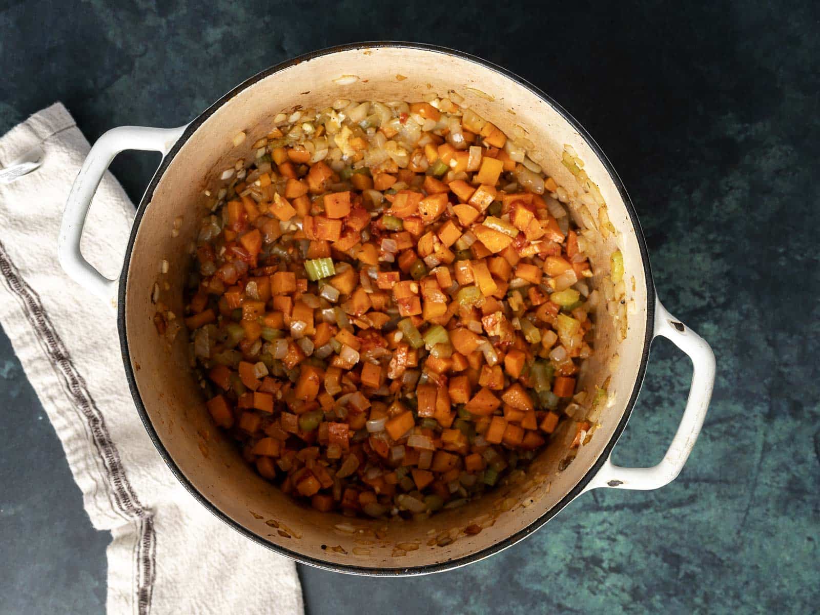 A pot filled with diced sweet potatoes, celery, and onions is transformed into a hearty ragu. A white cloth lies beside the pot on a dark green surface, setting the scene for this comforting dish.