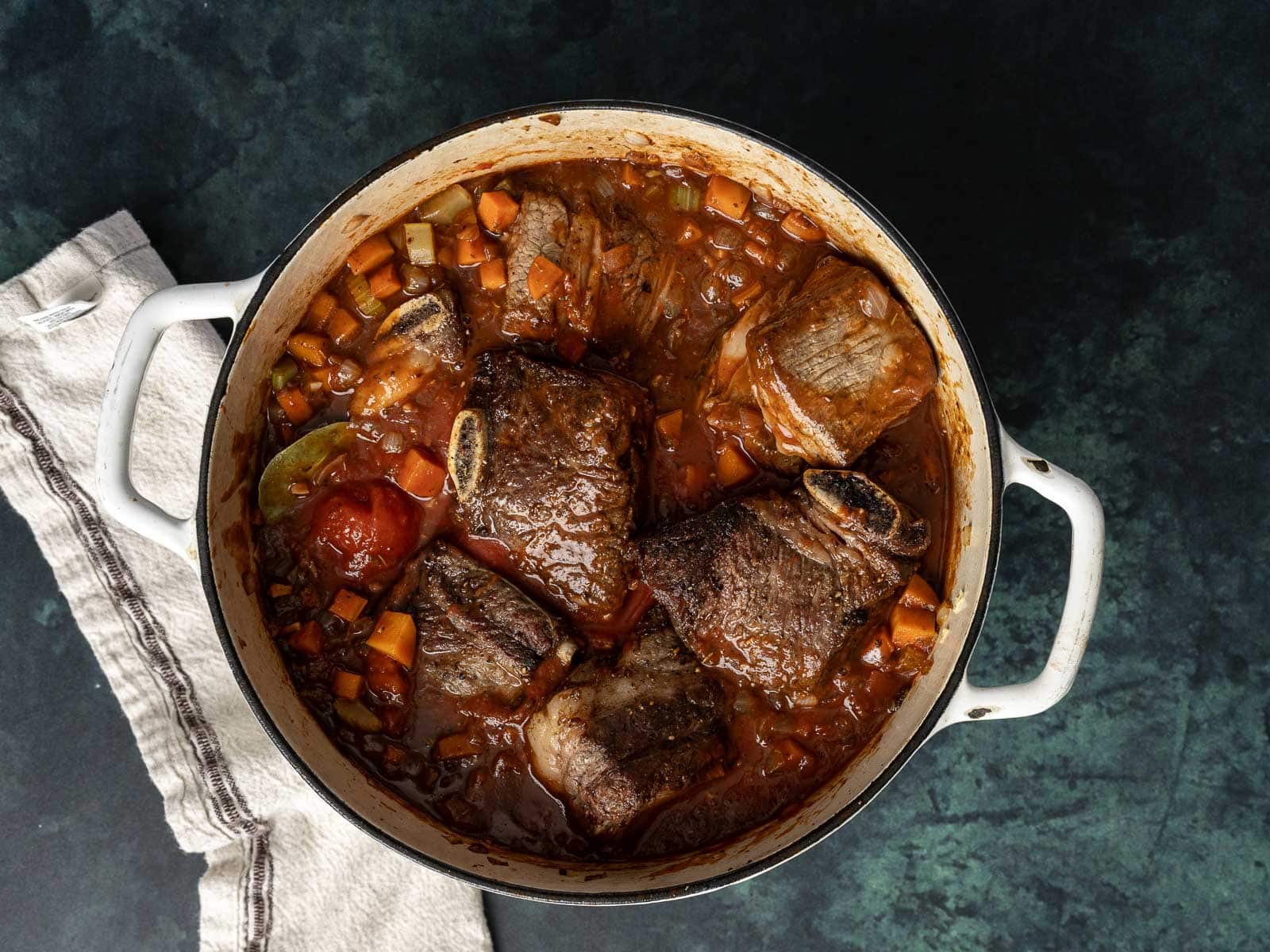 A pot of the best braised beef ragu in tomato sauce with carrots and herbs rests on a dark surface next to a white towel.