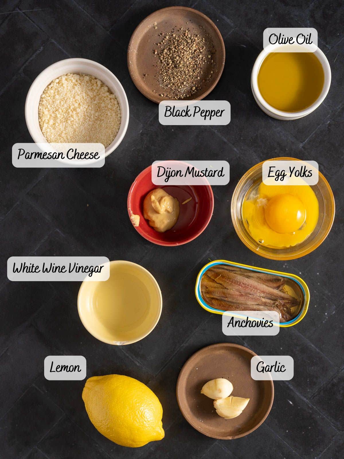 Ingredients for an anchovy Caesar dressing arranged on a dark surface: olive oil, black pepper, Parmesan cheese, Dijon mustard, egg yolks, savory anchovies, white wine vinegar, lemon, and garlic.