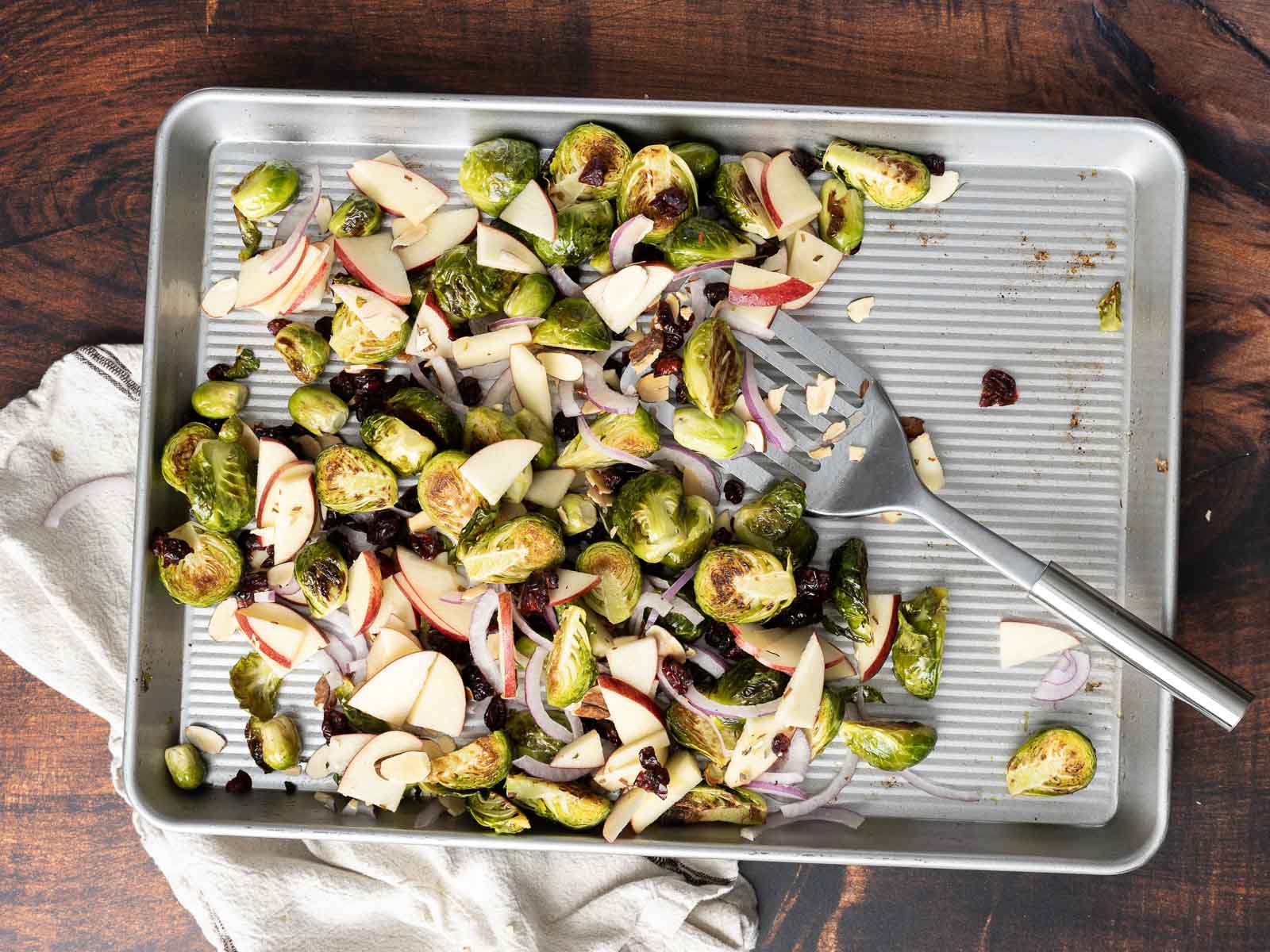 A baking tray features a roasted Brussels sprout salad with apple slices, onions, and cranberries, topped with a metal serving fork.