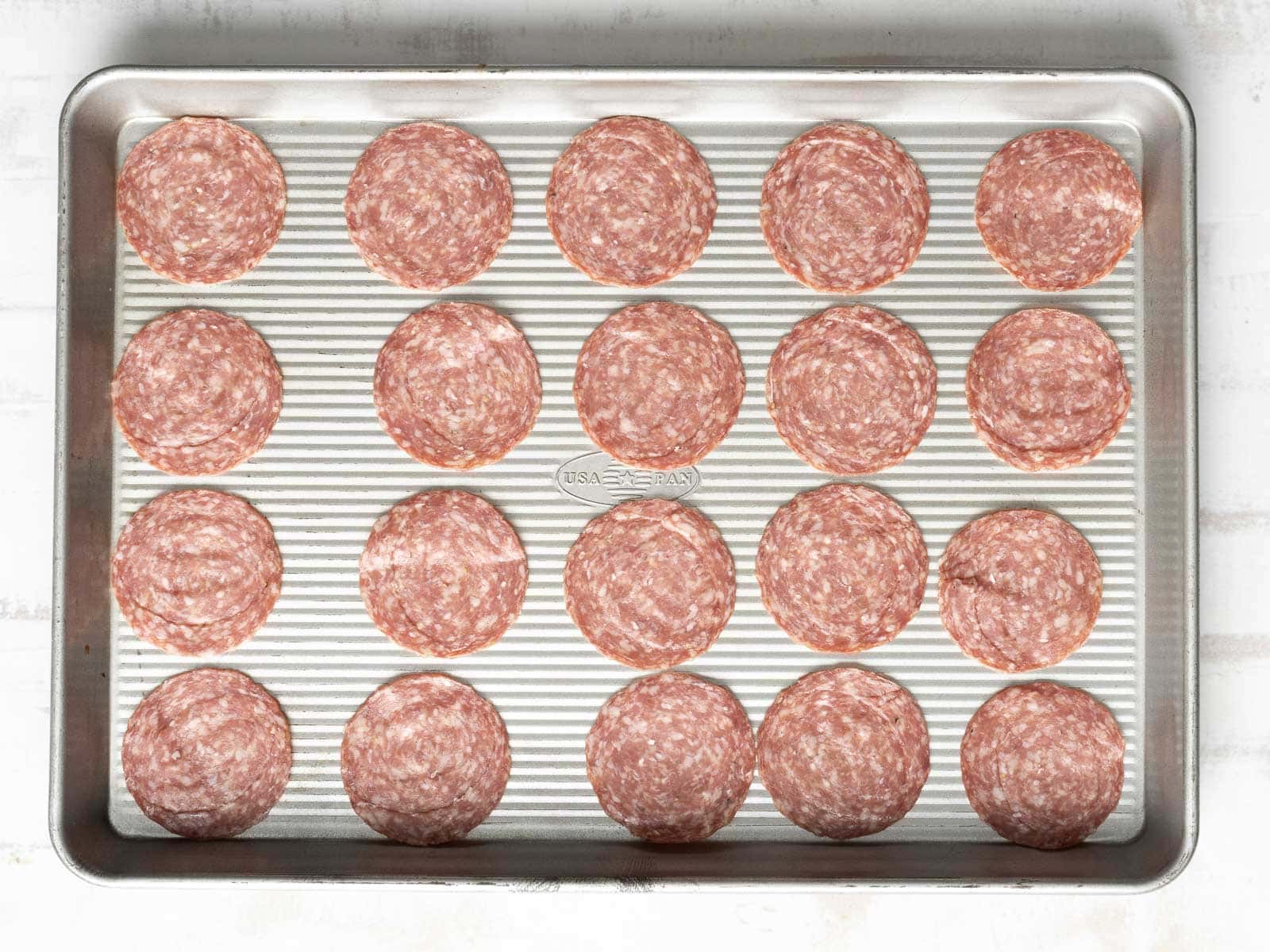 A baking sheet showcases four rows of five round, evenly spaced pepperoni slices, perfectly complemented by savory salami chips for an added crunch.