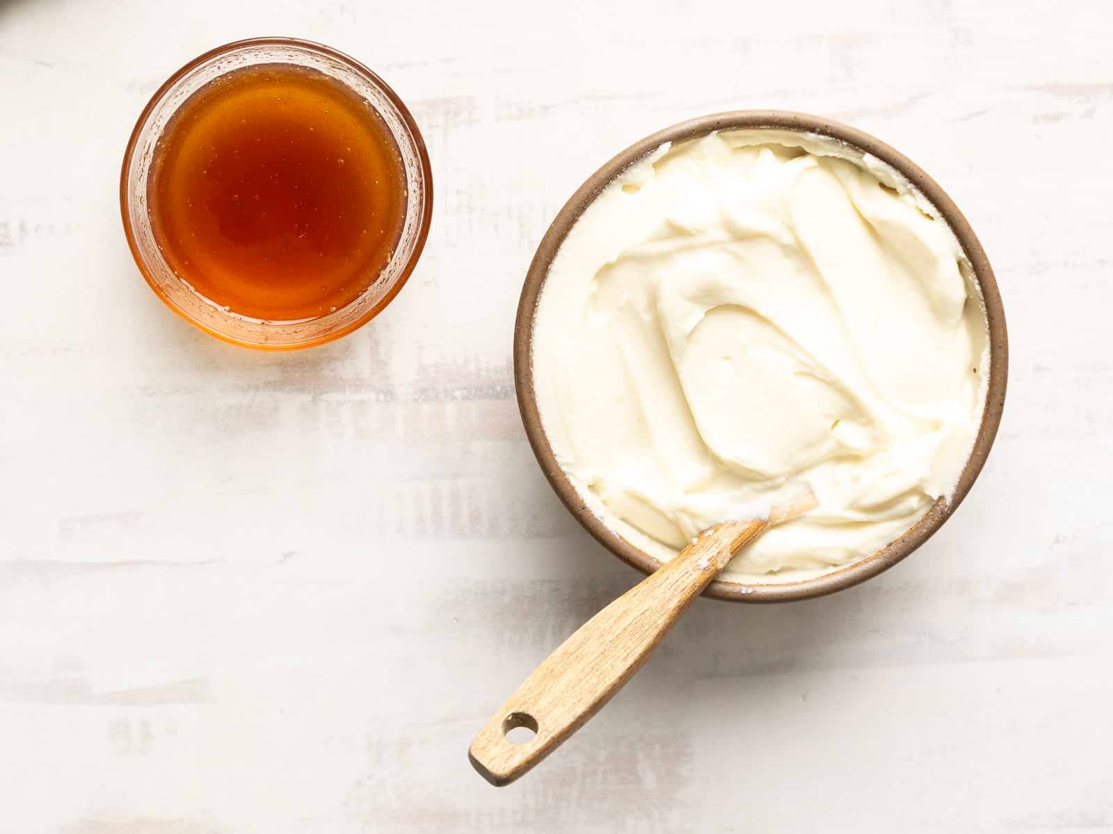 A bowl of creamy mascarpone cheese with a wooden spoon sits next to a small bowl of honey on a light surface, reminiscent of a delectable Honey Whipped Ricotta experience.