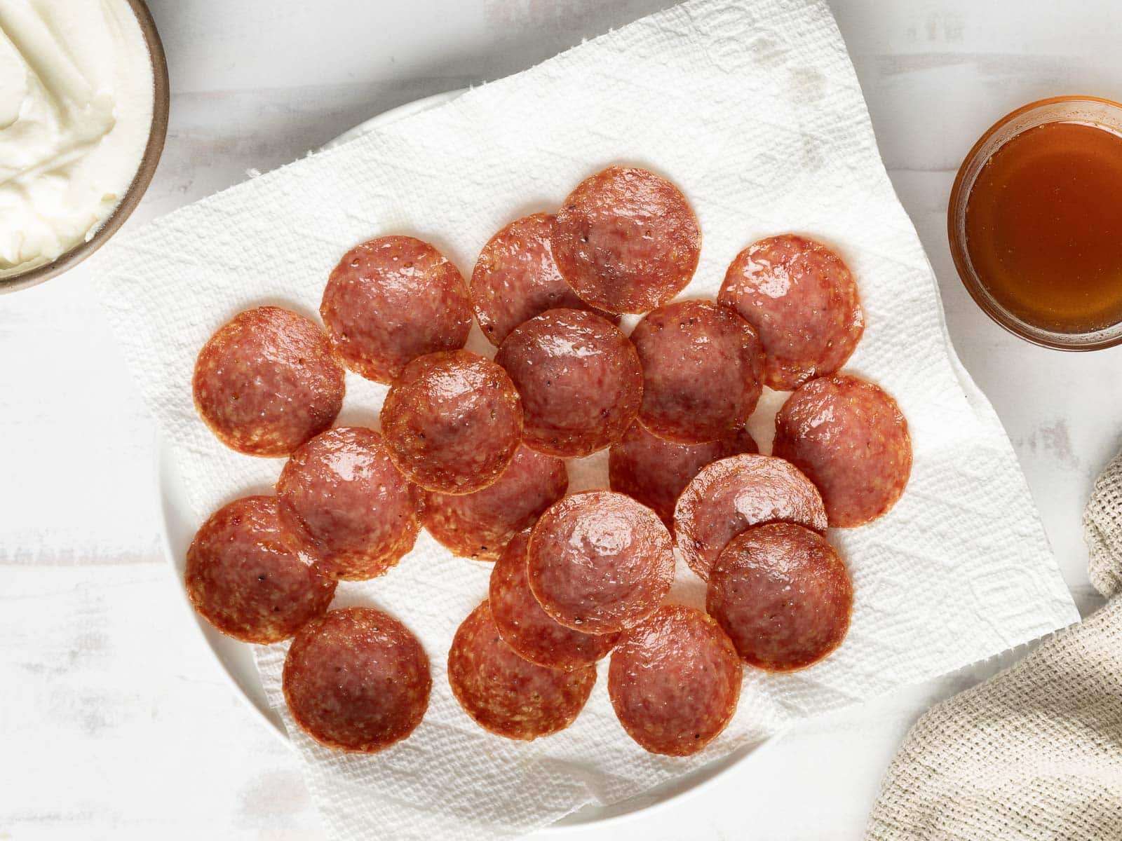 Sliced pepperoni on a paper towel-lined plate sits alongside a bowl of sauce and a bowl of ricotta cream on a pristine white surface.