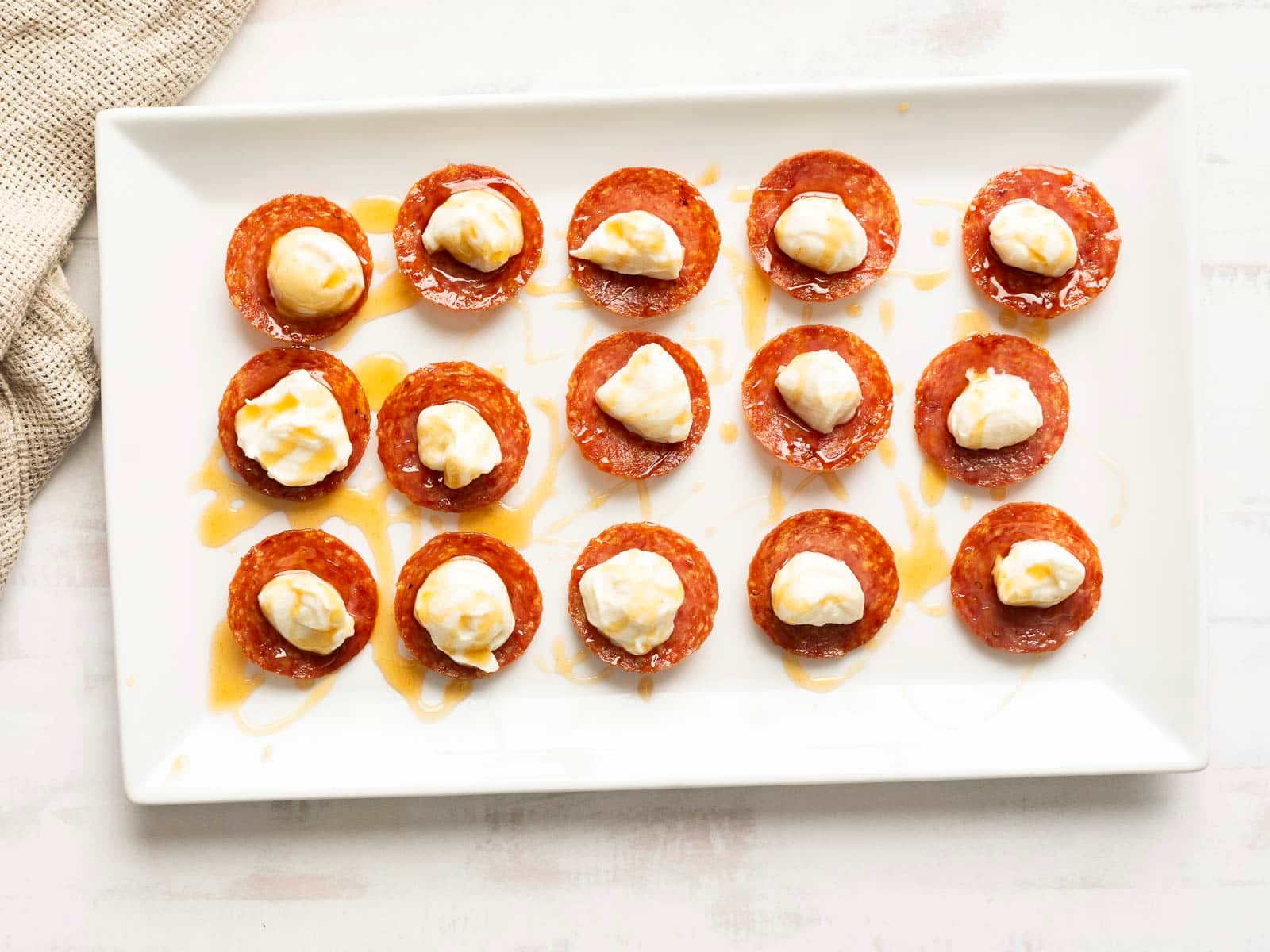 Sixteen slices of pepperoni topped with dollops of cream cheese and lightly accented by honey-whipped ricotta, elegantly arranged alongside crispy salami chips on a white rectangular plate.