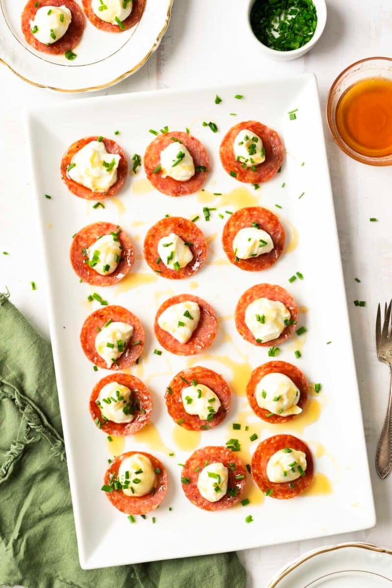 A platter of salami chips topped with honey whipped ricotta and garnished with chopped herbs.