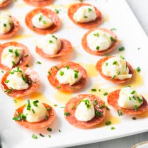 A close-up of pepperoni slices topped with scoops of honey whipped ricotta and garnished with chopped chives, all elegantly arranged on a white rectangular plate.