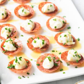 A close-up of pepperoni slices topped with scoops of honey whipped ricotta and garnished with chopped chives, all elegantly arranged on a white rectangular plate.
