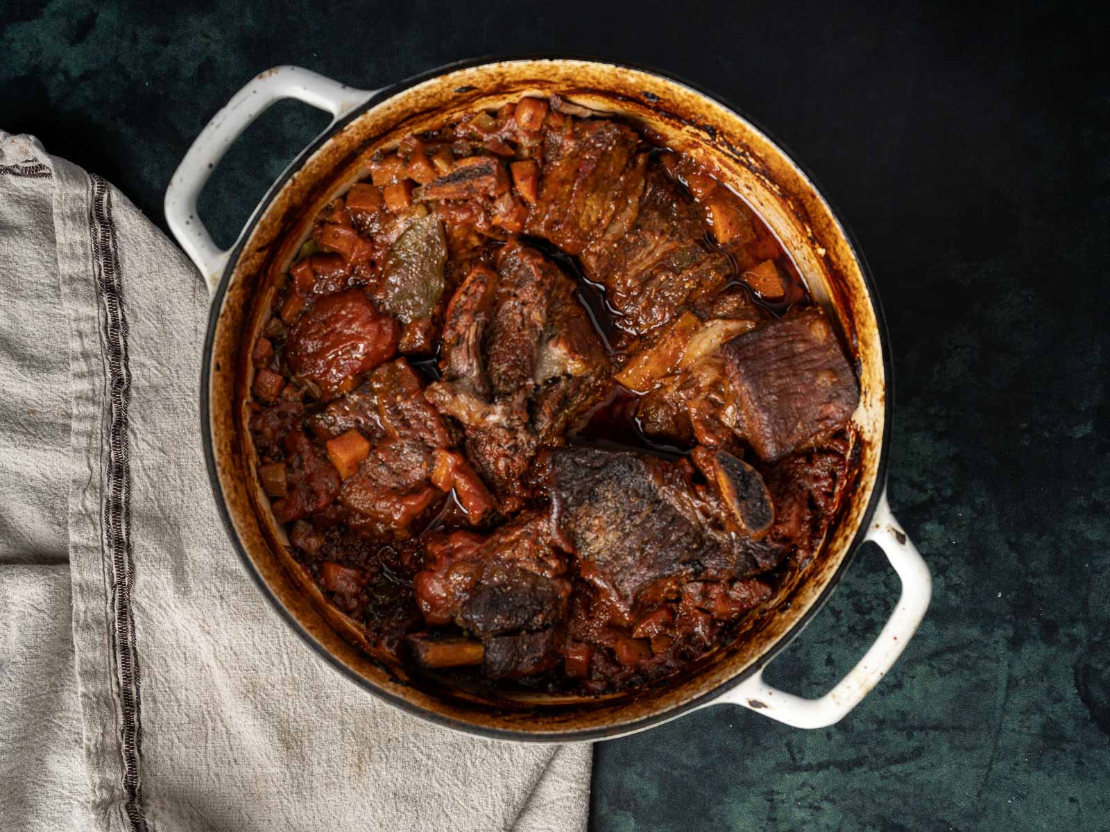 A pot of the best braised beef stew with vegetables rests on the dark surface, next to a neatly folded cloth.