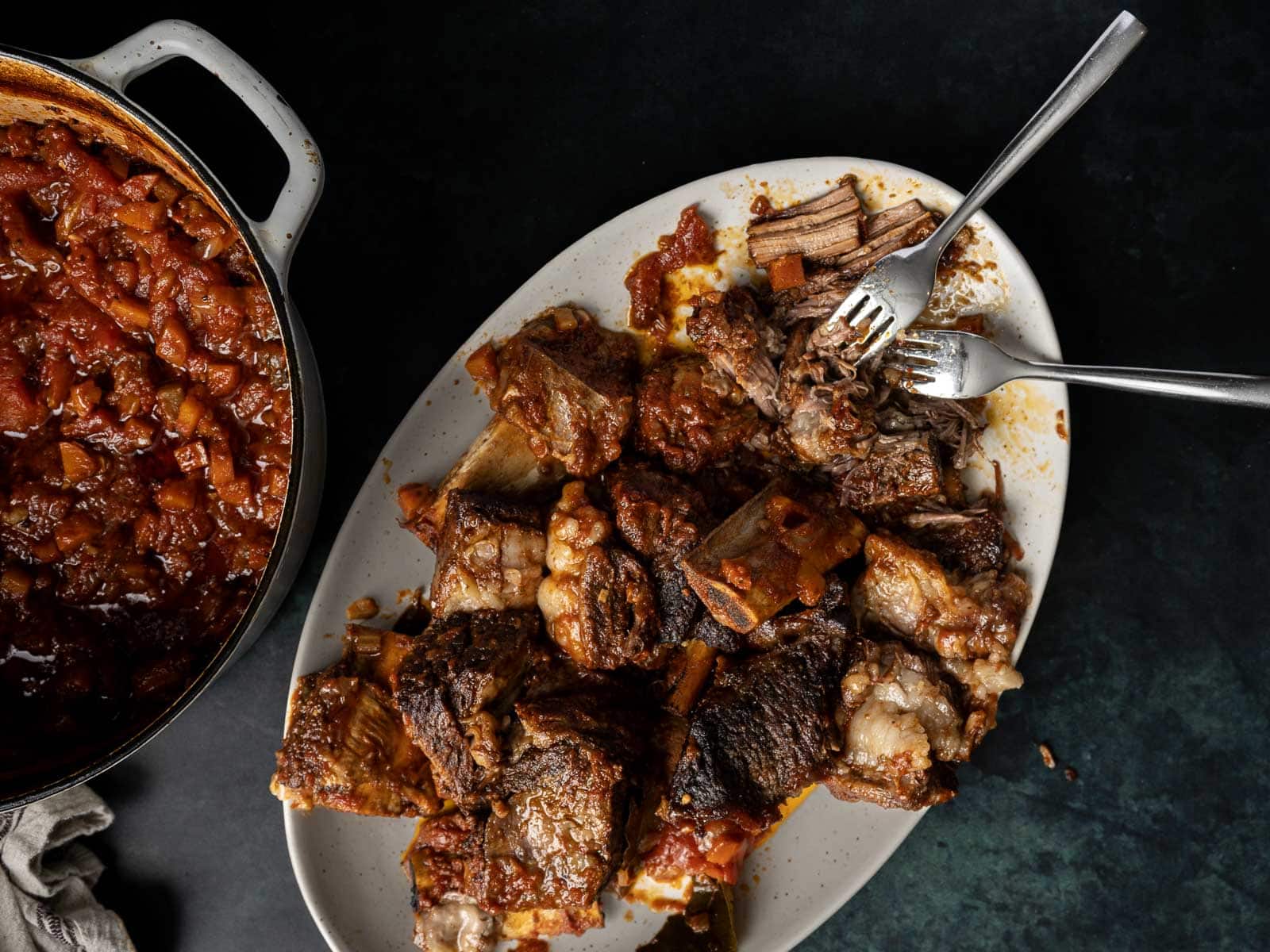 A dish of the best braised beef short ribs is served on a white plate next to a pot of cooked beans. Two forks are elegantly placed on the plate, inviting you to indulge in this tender, flavorful delight.