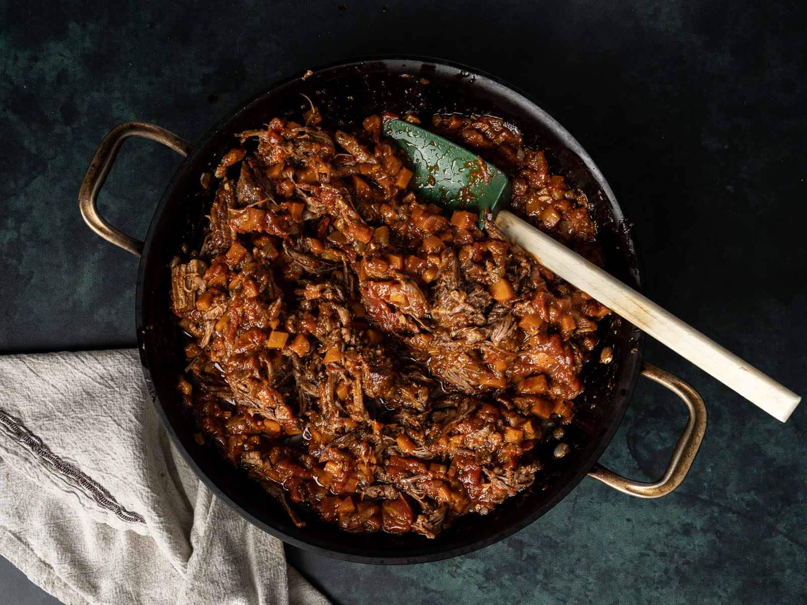 A black skillet brimming with braised beef ragu simmers in a rich, tomato-based sauce, while a green spatula and beige cloth rest beside the pan.