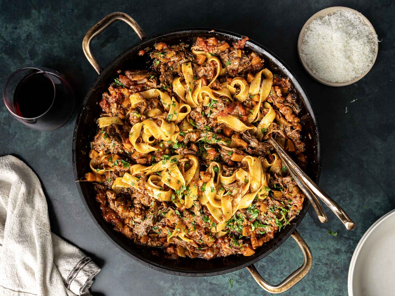 A skillet of pappardelle pasta with rich short rib ragu and herbs, alongside a glass of red wine, a bowl of grated cheese, a white napkin, and a white plate.