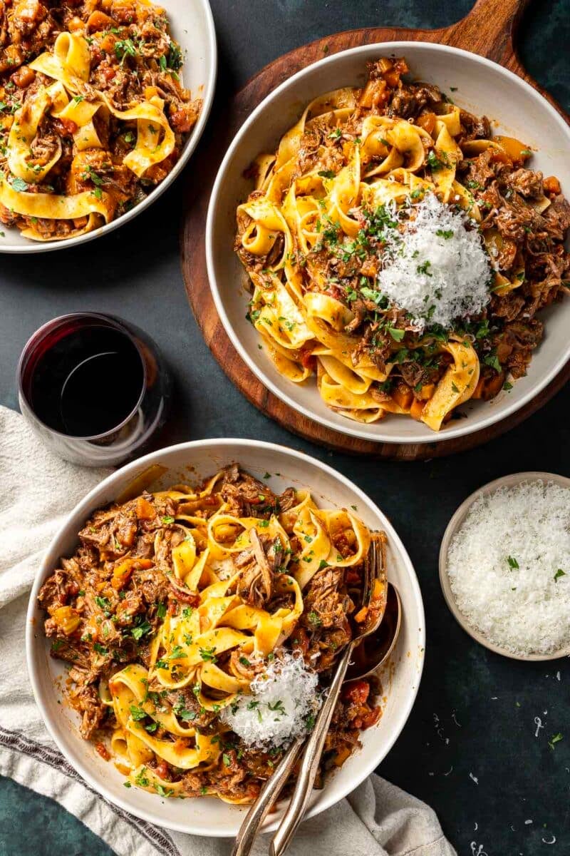 Three plates of pasta topped with rich ragu meat sauce and grated cheese, served alongside a glass of red wine and a bowl of additional cheese on the side.