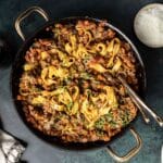 A pan of pasta with rich short rib ragu, garnished with fresh herbs. Forks rest inside the pan, inviting you in, while a small bowl of grated cheese waits nearby to complete this delectable experience.