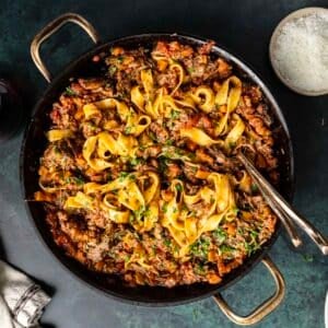 A pan of pasta with rich short rib ragu, garnished with fresh herbs. Forks rest inside the pan, inviting you in, while a small bowl of grated cheese waits nearby to complete this delectable experience.