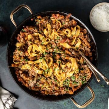 A pan of pasta with rich short rib ragu, garnished with fresh herbs. Forks rest inside the pan, inviting you in, while a small bowl of grated cheese waits nearby to complete this delectable experience.