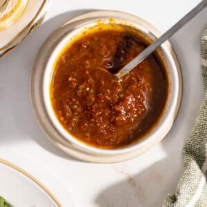 Top view of a bowl filled with thick, reddish-brown sun-dried tomato vinaigrette and a spoon resting inside. The bowl is on a light-colored surface with part of a green cloth visible nearby.
