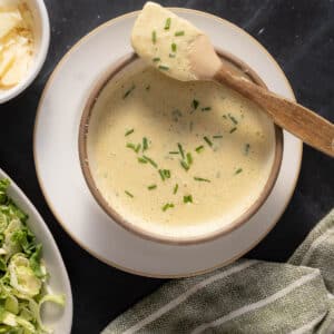 A bowl of creamy soup garnished with chopped chives, served with a wooden spoon. Visible in the background are a plate of leafy greens drizzled with anchovy Caesar dressing and a bowl of shredded cheese.