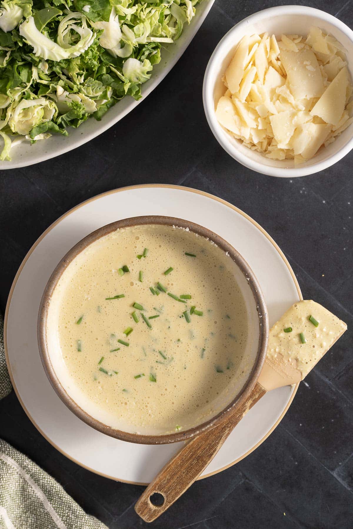 A bowl of creamy anchovy caesar dressing garnished with chopped chives sits alongside a parmesan cheese wedge. Nearby, a bowl of shaved parmesan and a plate of brussels sprouts complete the appetizing scene.